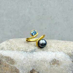 Geschwungener Ring mit oben offener Ringschiene aus 585/- Gelbgold mit einer dunkelgrau-schwarzen Tahitiperle (7.5-8.5mm) und einem ovalen Aquamarin ( ca. 5.5x5mm). Der Ring ist poliert und an den Seiten mit einer Wellenstruktur versehen.